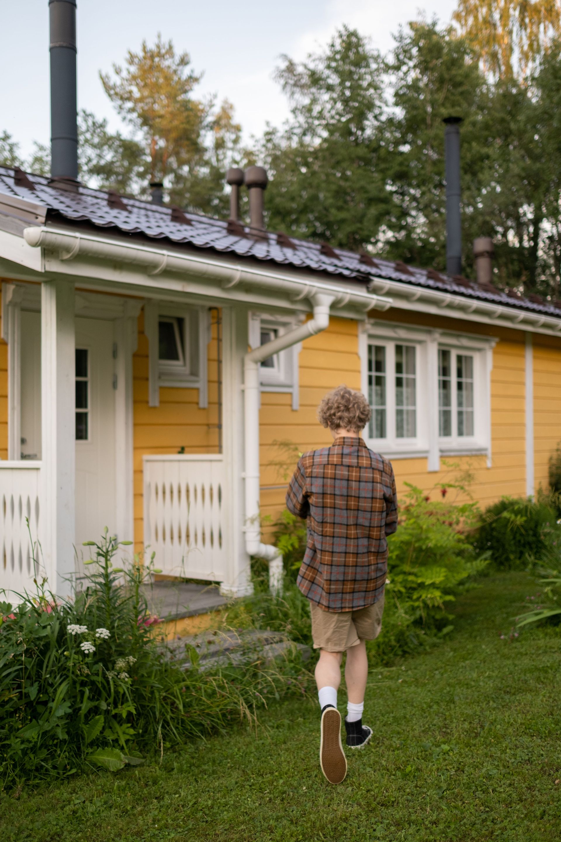 A teenager walks towards a serene yellow cottage surrounded by lush greenery on a summer day.